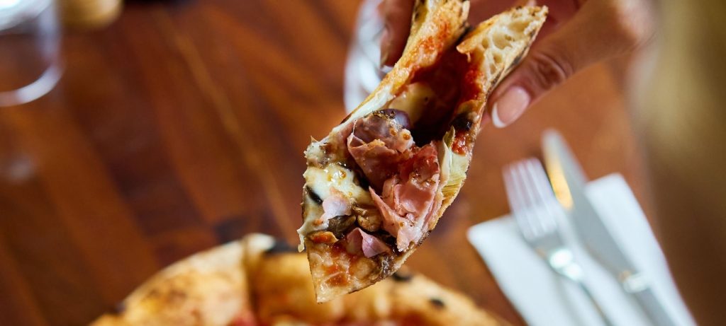 Hand lifting a slice of wood-fired prosciutto pizza at Il Verde Italian restaurant in Bowen Hills Brisbane